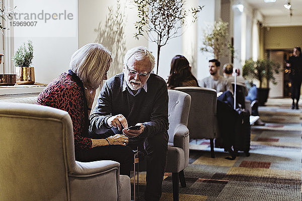 Älterer Mann zeigt einer Frau ein Smartphone  während er im Hotel auf einem Stuhl sitzt