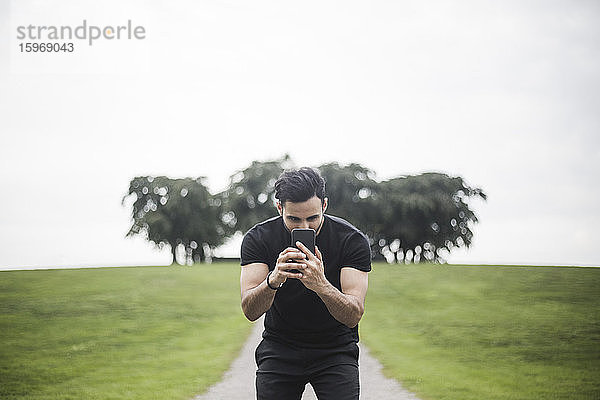 Junger Mann fotografiert durch ein Smartphone  während er auf einem Fußweg gegen den Himmel steht