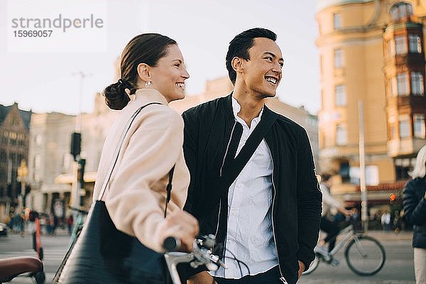 Lächelnde junge Frau mit Fahrrad schaut weg  während sie in der Stadt bei einem männlichen Freund steht