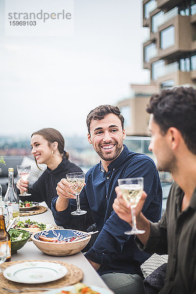 Lächelnder Mann unterhält sich mit einem Freund  während er während einer Party auf dem Dach neben einer Frau sitzt