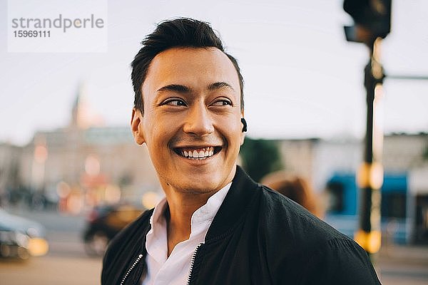 Lächelnder Mann schaut weg  während er in der Stadt In-Ohr-Kopfhörer trägt