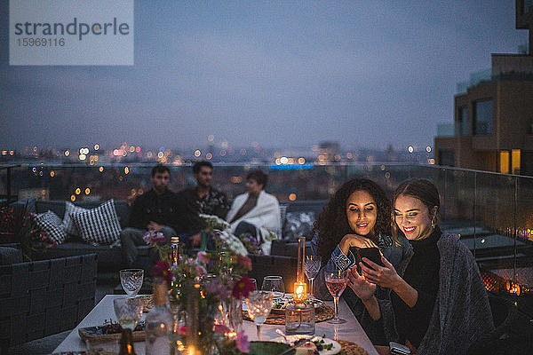 Lächelnde Frau zeigt einer Frau ein Smartphone  während Freunde im Hintergrund während eines geselligen Beisammenseins im Gebäude terr sitzen