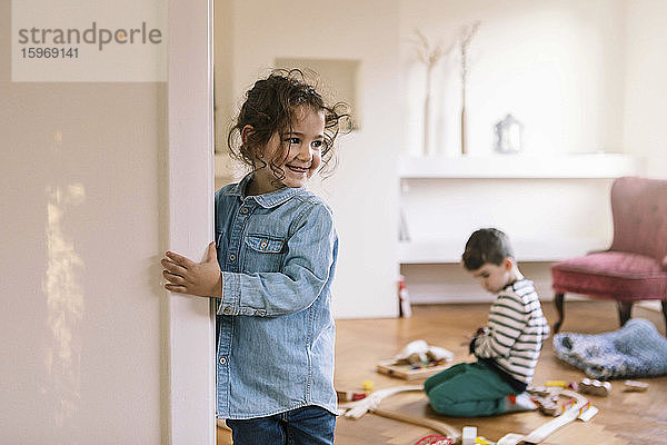 Lächelndes Mädchen steht an der Wand  während der Bruder zu Hause im Hintergrund spielt