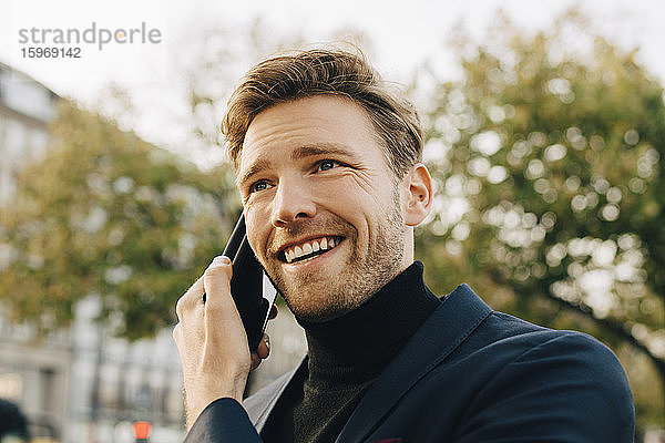 Lächelnder Geschäftsmann spricht durch ein Smartphone  während er in der Stadt wegschaut