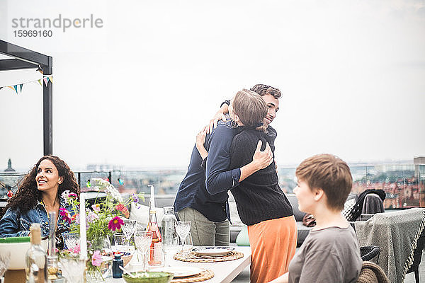 Junge männliche und weibliche Freunde umarmen sich  während sie an einem Tisch auf der Terrasse stehen