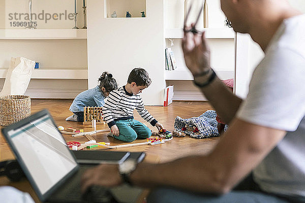 Vater mit Laptop schaut Kindern beim Spielen zu Hause zu