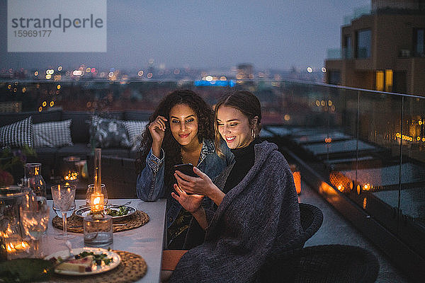 Lächelnde Frau zeigt einer Freundin ihr Smartphone während eines geselligen Beisammenseins auf der Terrasse des Gebäudes