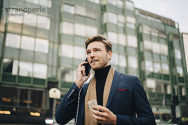 Selbstbewusster Geschäftsmann mit Wrap-Sandwich  der in der Stadt stehend per Smartphone spricht