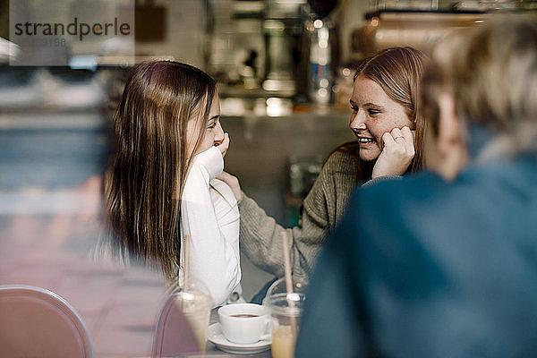 Lächelnde Teenager-Freundinnen unterhalten sich  während sie in einem Café sitzen und durch ein Glasfenster gesehen werden