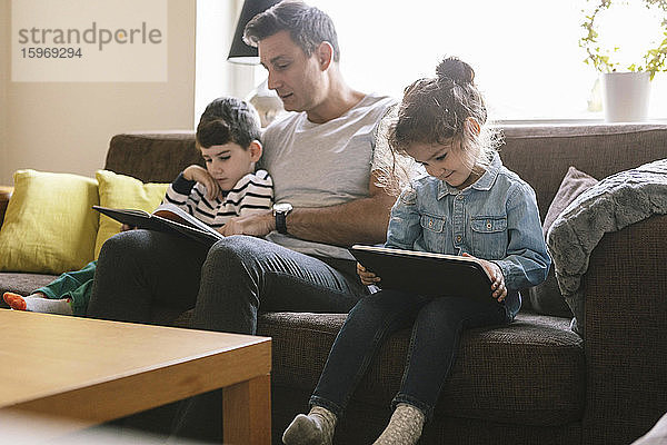 Vater liest dem Sohn ein Buch vor  während die Tochter ein digitales Tablett auf dem Sofa zu Hause benutzt