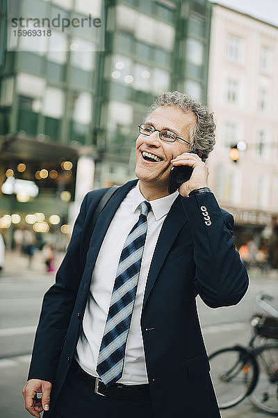 Lächelnder Geschäftsmann spricht durch ein Smartphone  während er in der Stadt steht