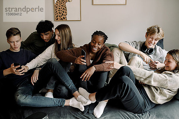 Lächelnde Teenager-Freunde nutzen soziale Medien auf Mobiltelefonen  während sie sich zu Hause auf dem Sofa entspannen
