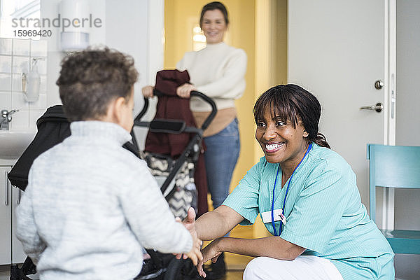 Lächelnde Krankenschwester schüttelt dem Jungen die Hand  während die Mutter im Hintergrund in der medizinischen Klinik steht