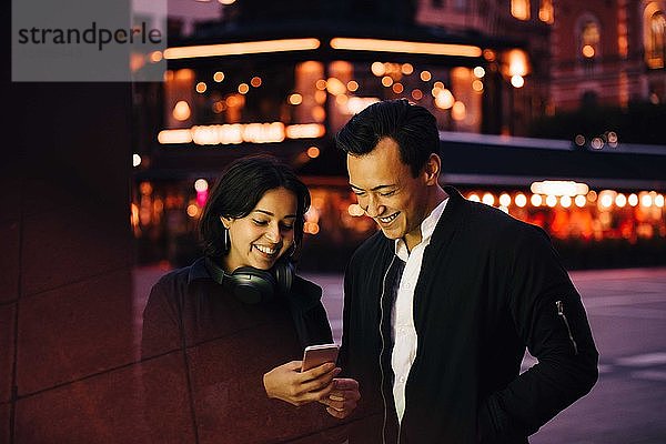Lächelnder Mann und lächelnde Frau  die ein Smartphone benutzen  während sie nachts in der Stadt stehen