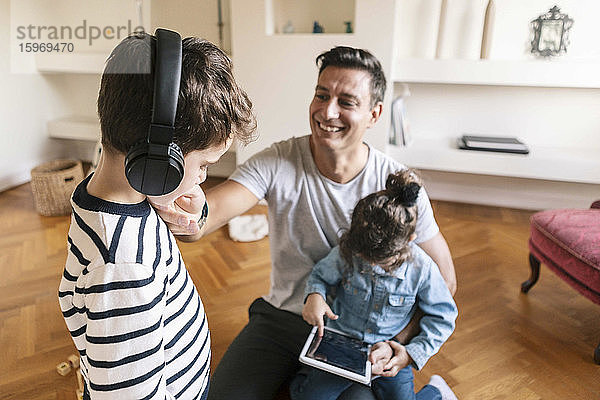 Lächelnder Vater sieht liebevollen Sohn an  während die Tochter zu Hause ein digitales Tablet benutzt