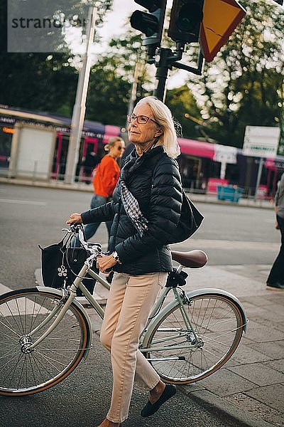 Ältere Frau mit Fahrrad überquerender Straße in der Stadt