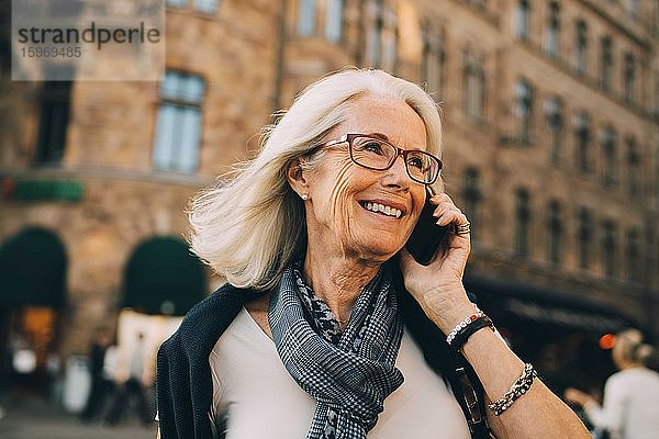 Lächelnde  runzlige Frau spricht am Telefon  während sie in der Stadt wegschaut