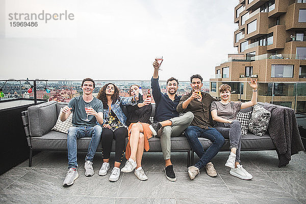 Porträt von lächelnden Freunden und Freundinnen mit Getränken  die auf dem Sofa auf der Gebäudeterrasse für geselliges Beisammensein sitzen