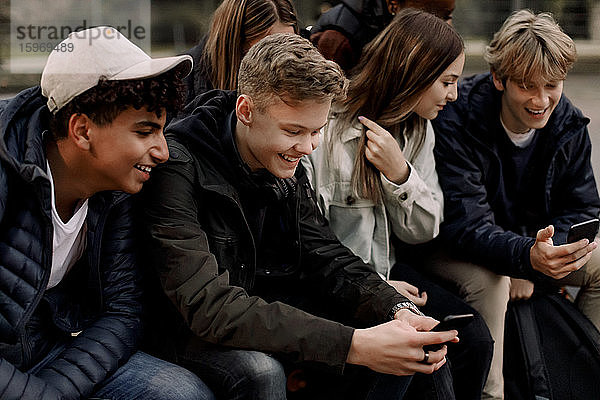 Lächelnde Mädchen und Jungen im Teenageralter benutzen Mobiltelefone  während sie im Park sitzen