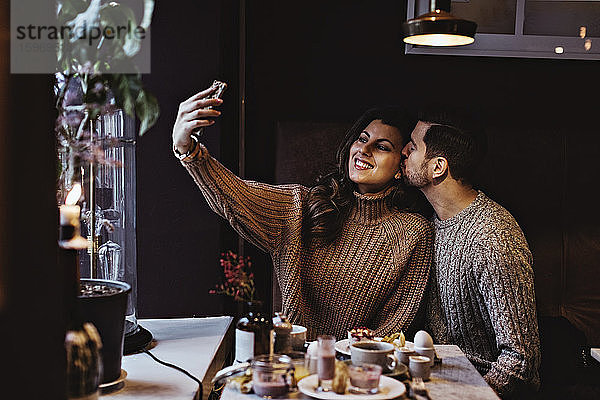 Lächelnde Frau  die sich über ein Smartphone selbstständig macht  während ihr männlicher Partner sich im Hotel küsst