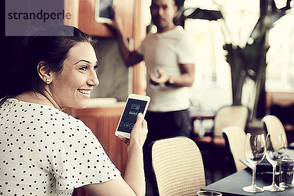 Rückansicht einer lächelnden Frau mit Smartphone  die wegschaut  während ein Mann im Cafe steht