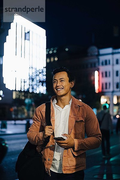 Lächelnder junger Mann schaut mit dem Telefon weg  während er nachts in einer beleuchteten Stadt steht