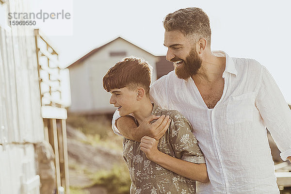 Lächelnder Vater umarmt Teenager-Sohn  während er am sonnigen Tag am Himmel steht