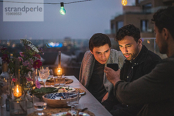 Mann zeigt männlichen Freunden ein Smartphone  während er bei geselligem Beisammensein auf der Terrasse sitzt