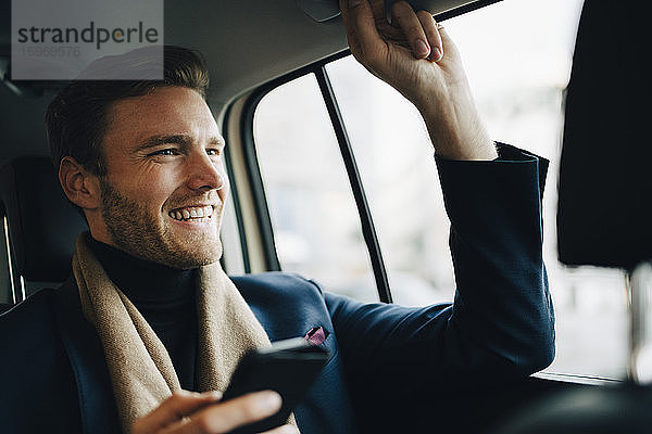 Lächelnder Geschäftsmann mit Telefon  der wegschaut  während er im Taxi sitzt