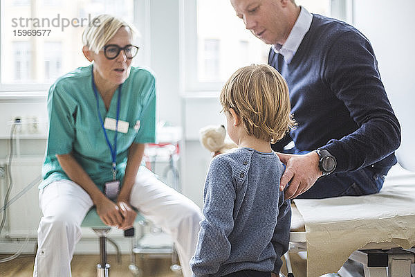 Rückansicht eines Jungen  der beim Vater steht  während er in der Klinik mit dem Arzt spricht