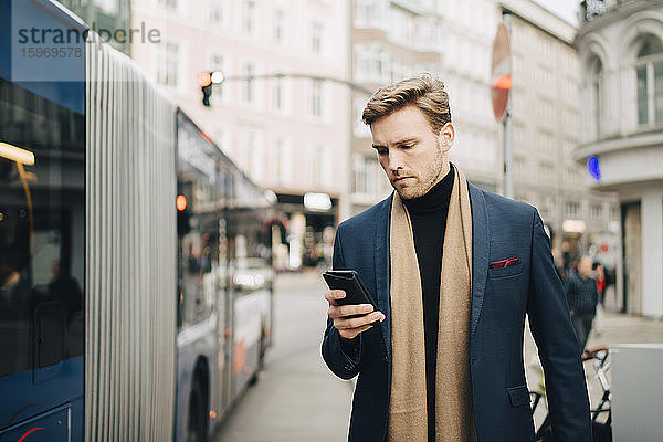 Männlicher Unternehmer nutzt Internet über Mobiltelefon  während er in der Stadt am Bus steht