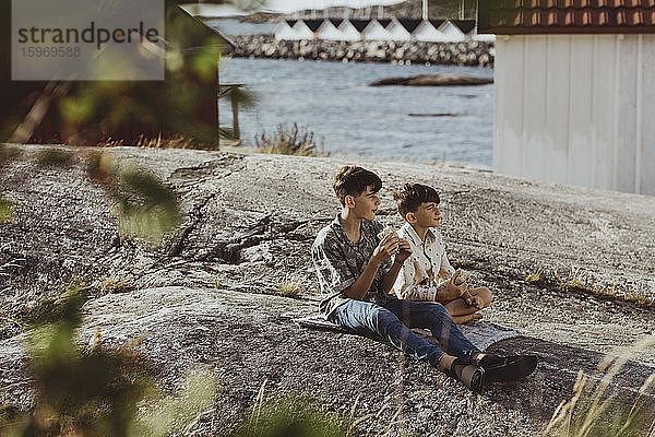 Männliche Geschwister schauen weg  während sie am Wochenende auf einer Decke über dem Archipel sitzen