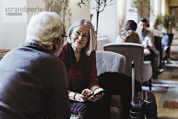 Ältere Frau mit Smartphone sieht Mann an  während sie im Hotel auf einem Stuhl sitzt