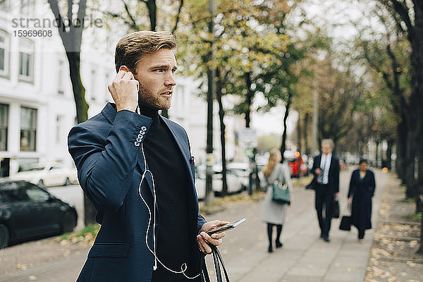 Geschäftsmann schaut weg  während er in der Stadt In-Ear-Kopfhörer hält