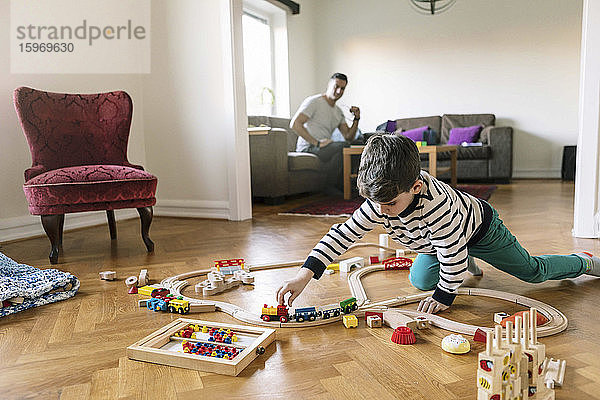Sohn spielt mit Miniatur-Eisenbahn  während der Vater zu Hause im Hintergrund auf dem Sofa sitzt