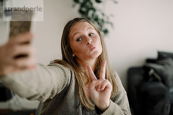 Teenager-Mädchen zeigt Friedenszeichen  während sie sich zu Hause über das Mobiltelefon selbstständig macht