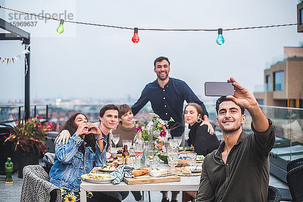 Lächelnder Mann  der sich über ein Smartphone selbstständig macht  während er sich mit Freunden in geselliger Runde auf der Terrasse des Gebäudes vergnügt
