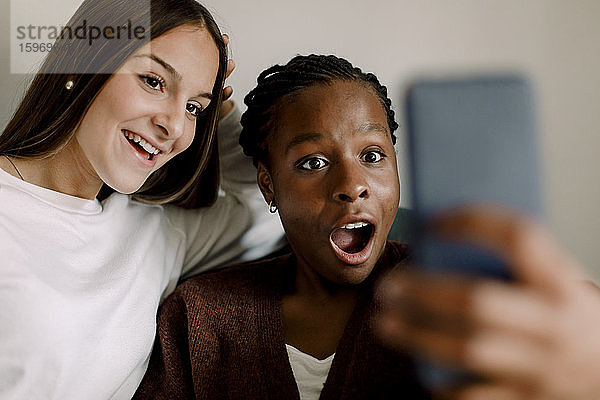Geschocktes Teenagermädchen schaut auf Smartphone  während es zu Hause beim lächelnden Freund Selfie sitzt