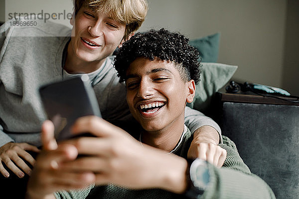 Lächelnder Teenager-Junge verschickt Textnachrichten über ein Smartphone  während er mit einem Freund zu Hause sitzt