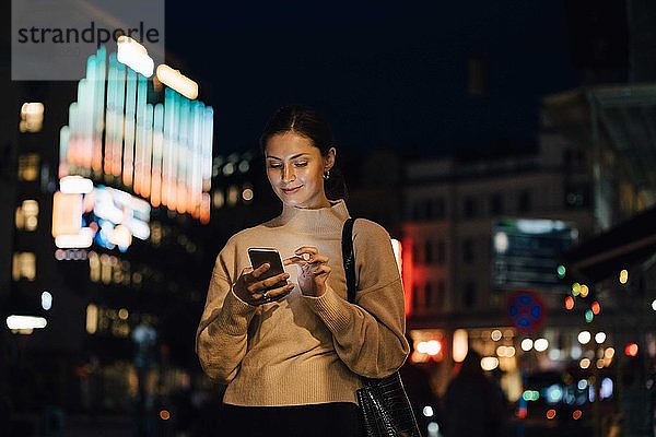 Lächelnde Frau schreibt nachts in der Stadt per Smartphone SMS