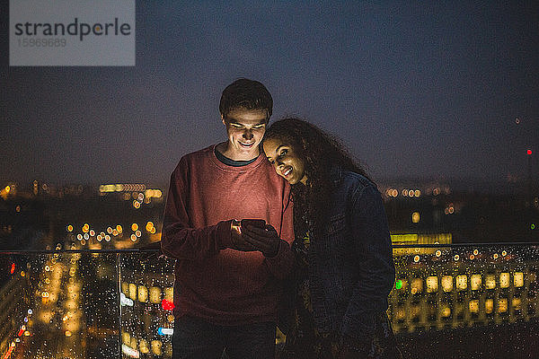 Junger Mann zeigt einer Freundin ein Smartphone während eines geselligen Beisammenseins auf einer Terrasse in der Nacht