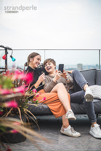 Fröhliche Frau benutzt Smartphone  während sie beim geselligen Beisammensein mit einem fröhlichen Freund auf dem Sofa sitzt