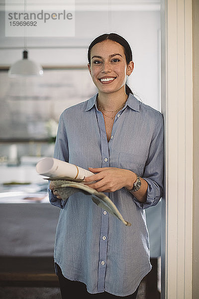 Portrait eines lächelnden Designprofis mit Papier- und Stoffmustern im Heimbüro stehend