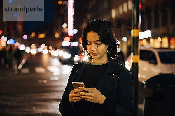 Junge Frau benutzt Mobiltelefon  während sie nachts in der Stadt steht