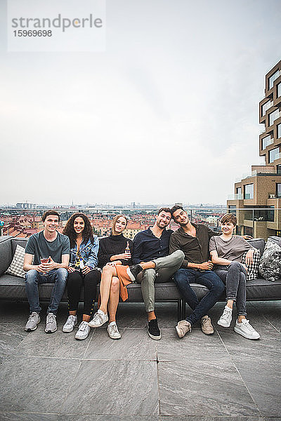 Porträt von lächelnden Freundinnen und Freunden  die auf einem Sofa auf einer Gebäudeterrasse für geselliges Beisammensein sitzen