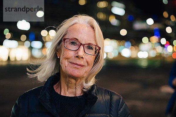 Porträt einer faltigen  lächelnden Frau  die nachts in einer beleuchteten Stadt steht