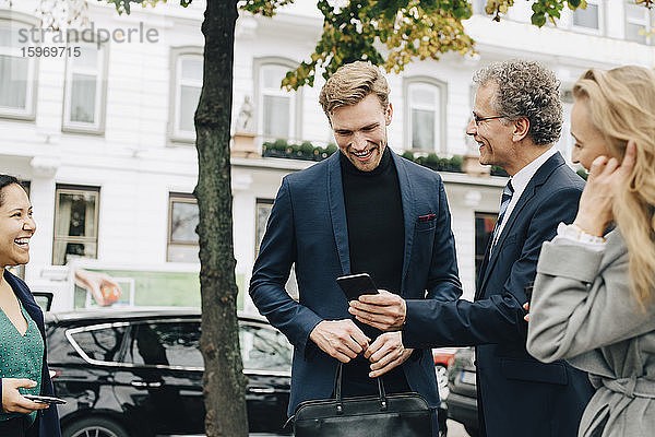 Lächelnder Unternehmer zeigt einem männlichen Kollegen sein Handy  während er in der Stadt bei seinen Kollegen steht