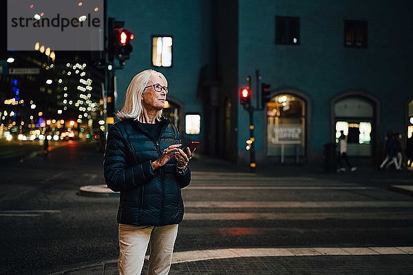 Lächelnde Frau mit Smartphone beim Aufblicken in beleuchteter Stadt bei Nacht