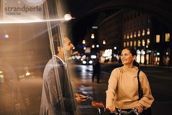 Lächelnde junge Frau mit Fahrrad schaut einen männlichen Freund an  während sie nachts in der Stadt steht