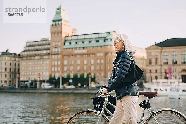 Seitenansicht einer lächelnden älteren Frau mit Fahrrad beim Überqueren eines Flusses in der Stadt